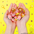Hands holding small character figurines against a yellow background with cartoonish decorations.
