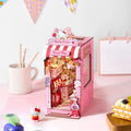 A pink Hello Kitty-themed miniature cardboard shop model displayed on a light pink tabletop, surrounded by cute desk props including a white mug with chopsticks, a small Hello Kitty figurine, and a slice of cake.