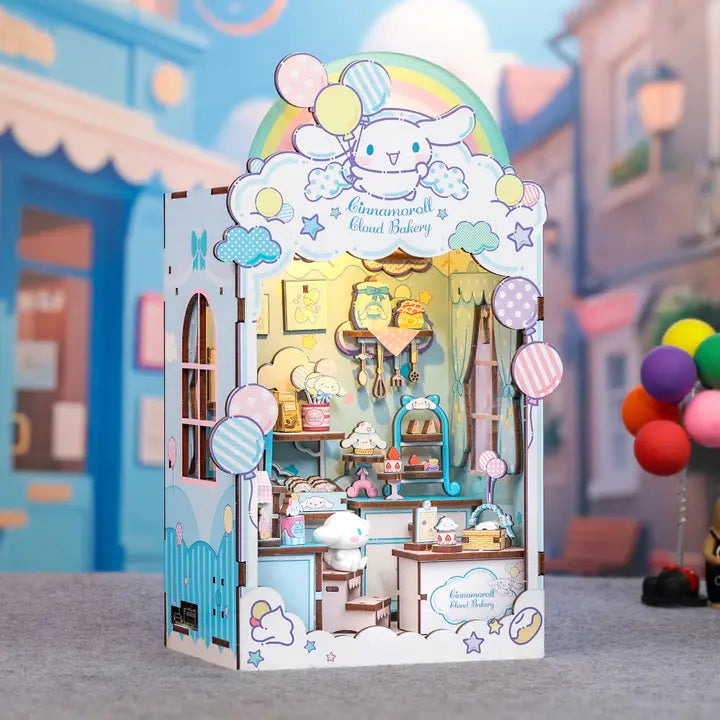 A 3D cardboard puzzle box of the character Cinnamoroll running a bakery, displayed on a gray surface with a blurred street scene background. The box is decorated with pastel colors, balloons, and features a detailed miniature bakery interior.