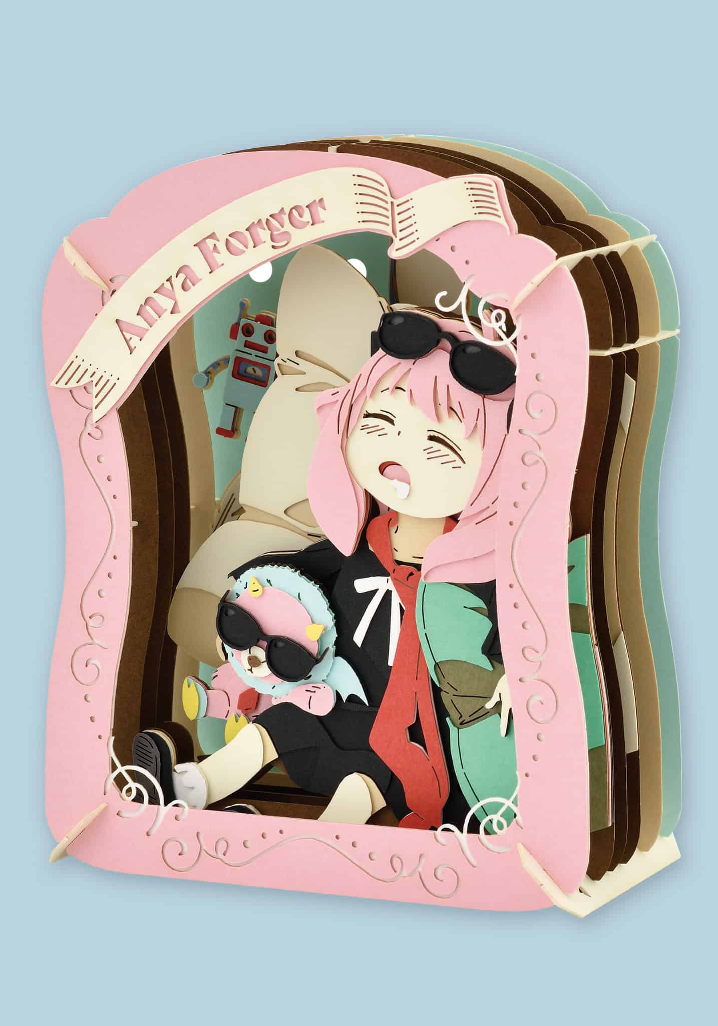 A 3D papercraft art piece depicting the anime character Aria Forger sitting inside a pink book frame, with 'Aria Forger' written on a banner at the top, set against a solid light blue background.