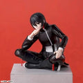 An anime figurine of character with black hair, wearing a black and white outfit, sitting cross-legged on a gray pedestal against a solid red background.
