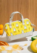 A yellow and white Miffy-patterned lunch bag with white handles, shown floating above a tabletop with fruit and a checkered cloth in a styled setting.