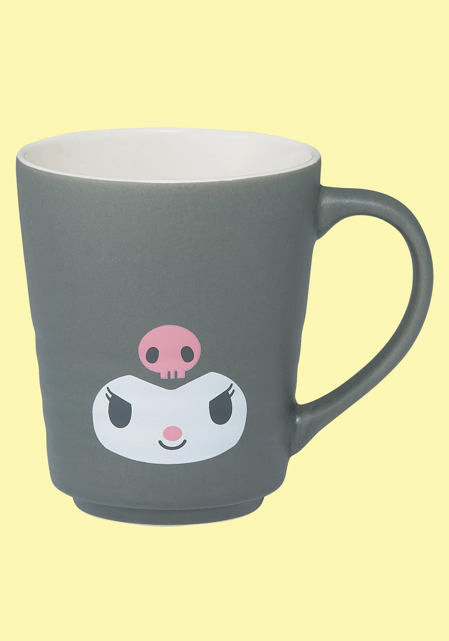 A dark gray ceramic mug with a white interior, featuring a cartoon character face printed on the front with a pink skull accent, set against a solid pale yellow background.
