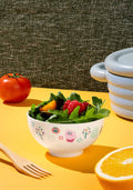 Bowl of salad with a floral pattern on a yellow surface, surrounded by a tomato, wooden fork, and half an orange.