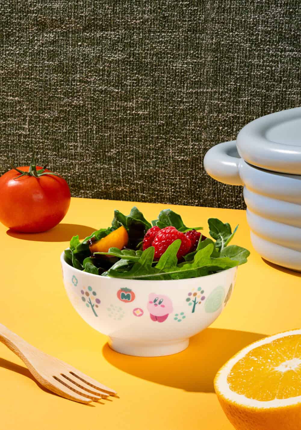 Bowl of salad with a floral pattern on a yellow surface, surrounded by a tomato, wooden fork, and half an orange.