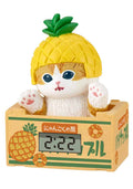 A cute digital alarm clock shaped like a white cat wearing a yellow pineapple hat, sitting inside a pineapple-themed box. The cat has blue eyes and pink paws, and the box features green and orange Japanese text and illustrations.