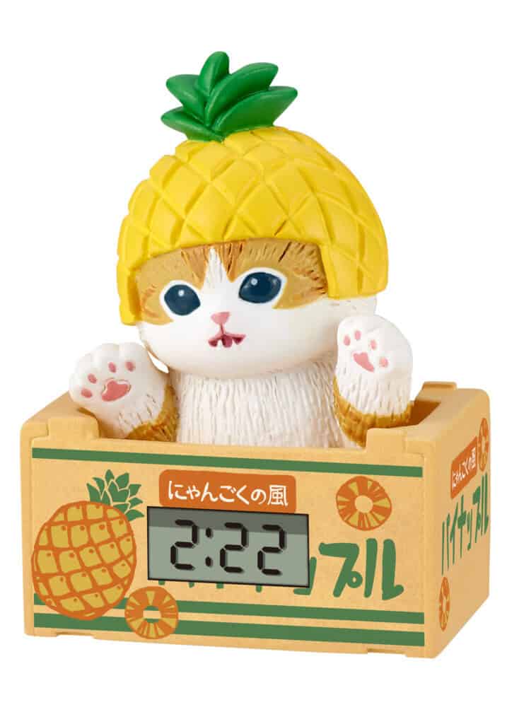 A cute digital alarm clock shaped like a white cat wearing a yellow pineapple hat, sitting inside a pineapple-themed box. The cat has blue eyes and pink paws, and the box features green and orange Japanese text and illustrations.