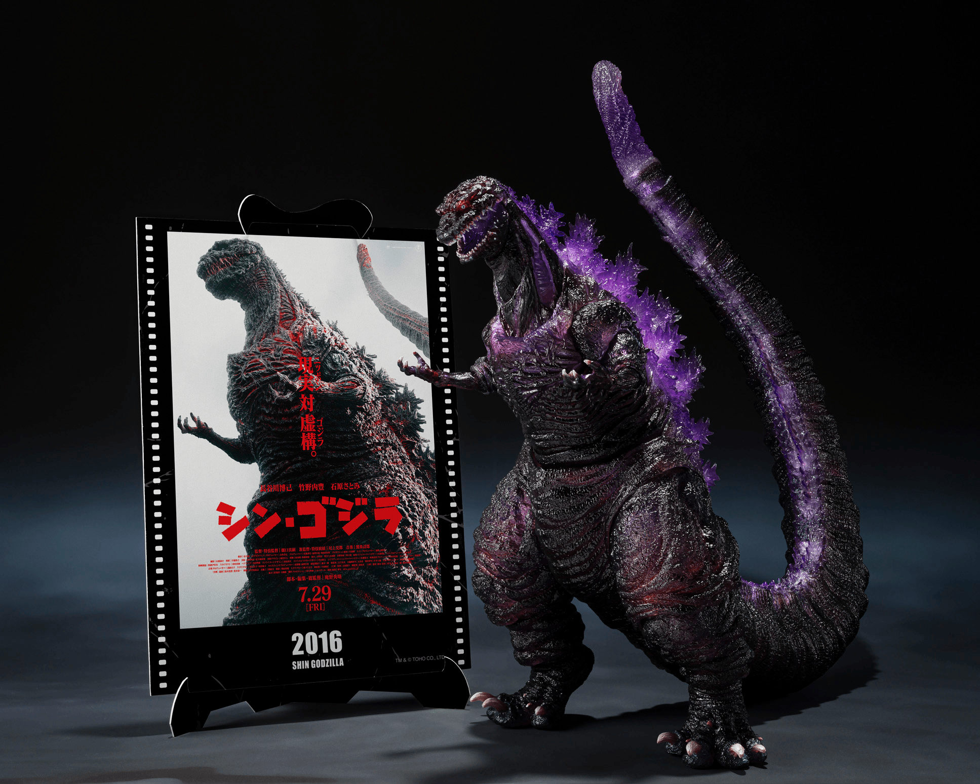 A Godzilla collectible figure stands beside a vertical Godzilla movie poster mounted on a small display stand, against a dark studio background.