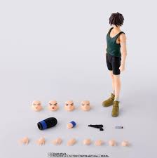 A small action figure or collectible character standing on a plain light gray background, with multiple interchangeable heads, hands, and small accessories arranged below it. The figure wears a dark green sleeveless top, black shorts, and olive-green shoes.