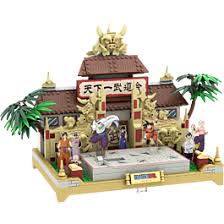 A small brick-built temple or shrine model with a brown roof, gold decorative details, a central gate, figures in front, palm trees, and a light wood base, shown on a plain white background.