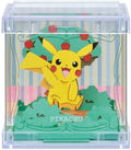 A clear acrylic display case containing a 3D papercraft of Pikachu sitting in front of a stylized green tree with red apples. The base features a green textured pattern with the word 'PIKACHU' printed on it.