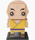 A small brick-built character figure with a bald head, black eyes, and a yellow robe with a purple stripe, standing on a black display base against a light gray background.