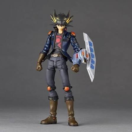 A detailed action figure of Yu-Gi-Oh! Yugi, standing on a gray studio background. He is wearing his signature blue jacket, gray pants, and brown boots, holding a white and blue card shield.